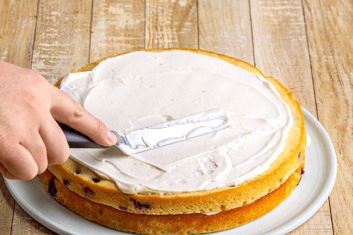 3/4th shot of a hand is seen spreading frosting onto a two layer cake using a spatula; the cake appears to be on a circular white plate placed on a wooden surface;