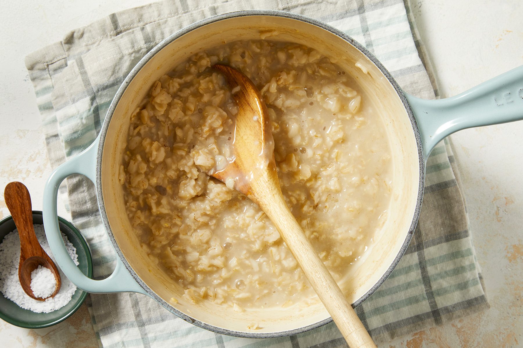 A pot of creamy oatmeal with a wooden spoon inside, placed on a striped cloth. A small bowl with salt and a wooden spoon is beside the pot.