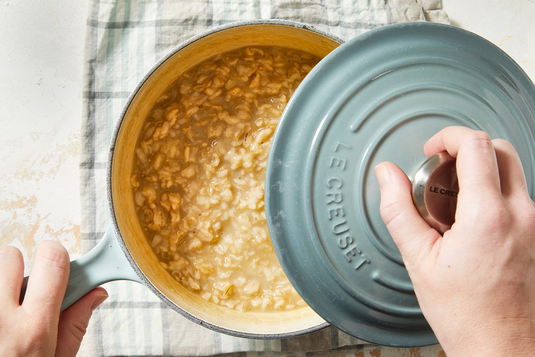 A person lifts the lid of a teal Le Creuset pot, revealing bubbling risotto inside. The pot rests on a checkered cloth on a light countertop.
