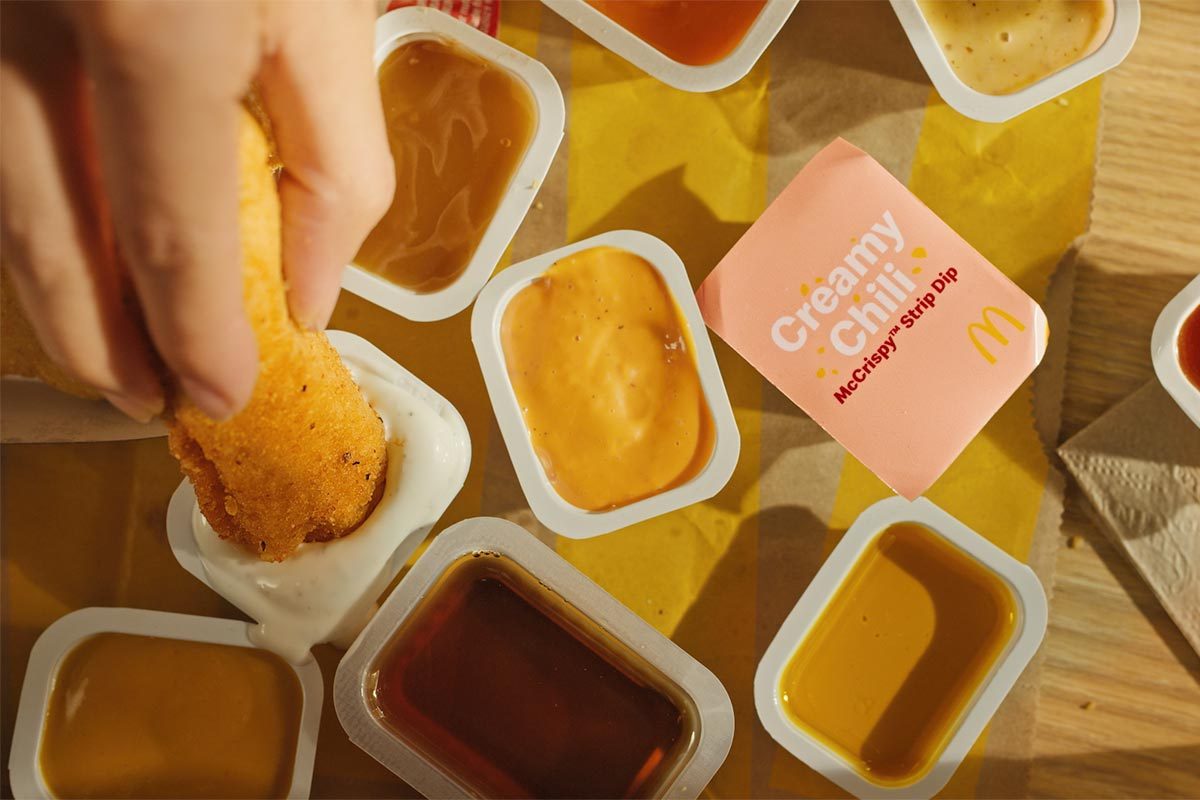 Birds eye angle of a hand dipping chicken strip into one of several packets of different colored sauces