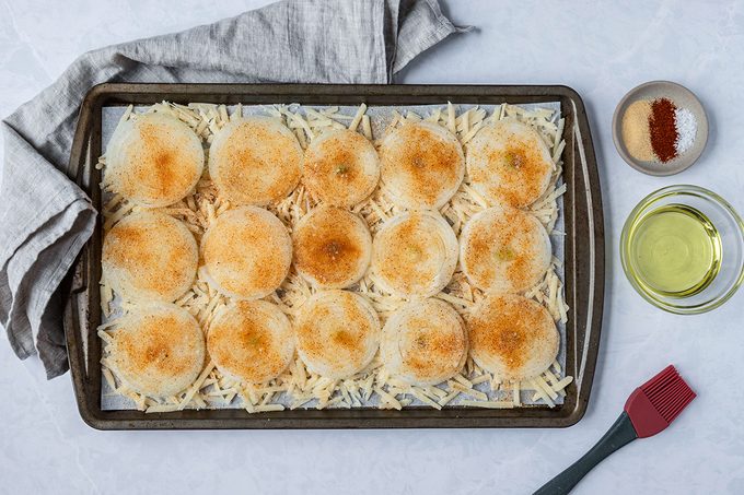 Round pastry circles topped with brown seasoning on a lined baking sheet, surrounded by shredded cheese. Nearby are bowls of oil and spices, and a red brush. A gray cloth is placed beside the tray on a white surface.