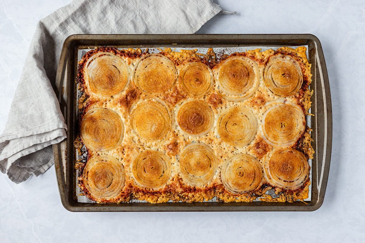 A baking sheet topped with golden-brown circular slices of bread, each surrounded by a crispy cheese crust. A light-colored cloth is draped over one corner of the sheet.