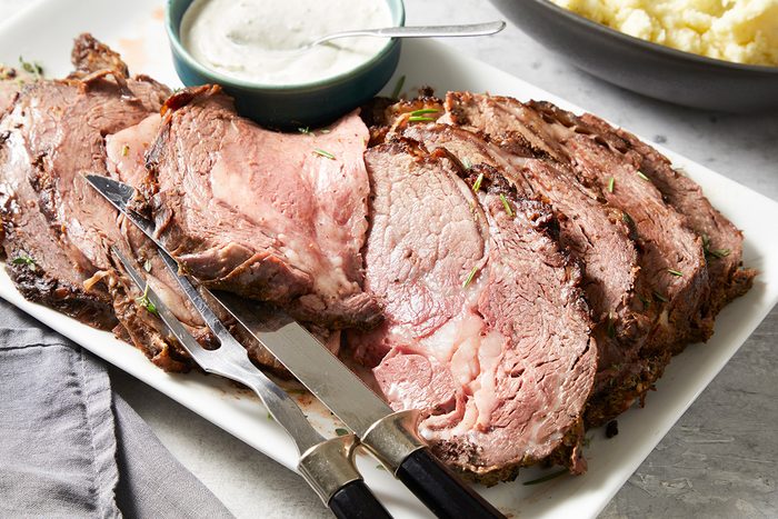 A sliced roast beef arranged on a rectangular white platter with a serving fork and knife. A small bowl of creamy sauce sits beside the meat. A bowl of mashed potatoes is partially visible in the background.