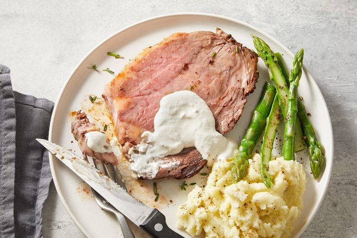 A plate with a slice of roast beef topped with creamy sauce, served with mashed potatoes and steamed asparagus. A knife and fork are placed beside the plate on a light gray surface.