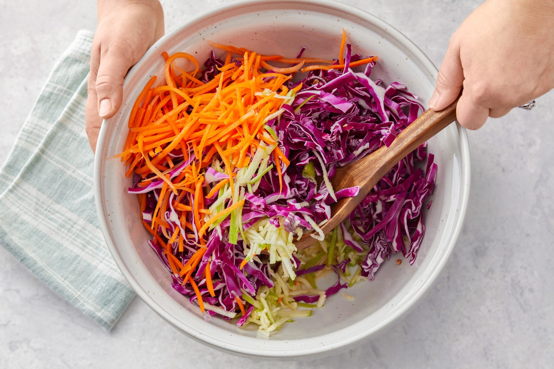 overhead shot of a large white bowl, Stiring in cabbage, apple, carrot and pecans with wooden spoon, a light colored kitchen cloth is visible