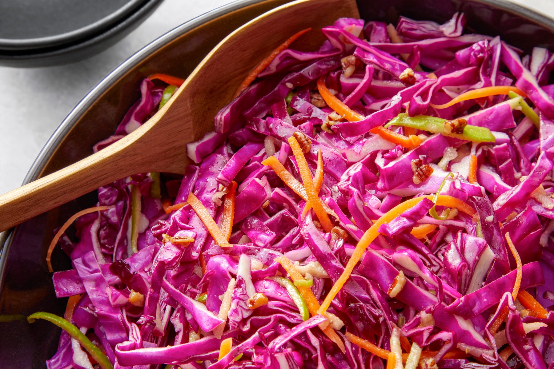 closeup shot of Red Cabbage Salad in a large bowl, a wooden serving spoon is placed into dish, dark colored bowl is slightly visible in background
