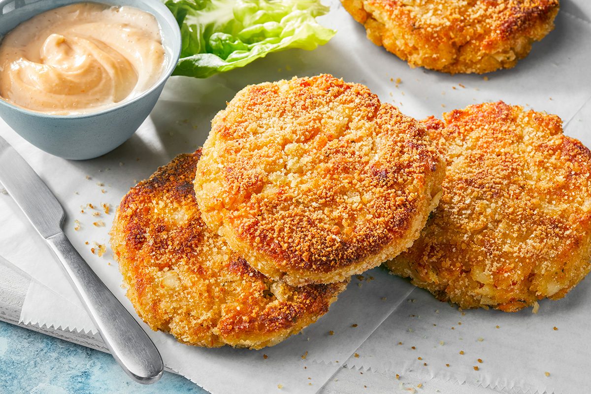 Three golden, breaded patties placed on parchment paper next to a small bowl of creamy dipping sauce and some fresh lettuce leaves. A knife and fork are partially visible on the side.