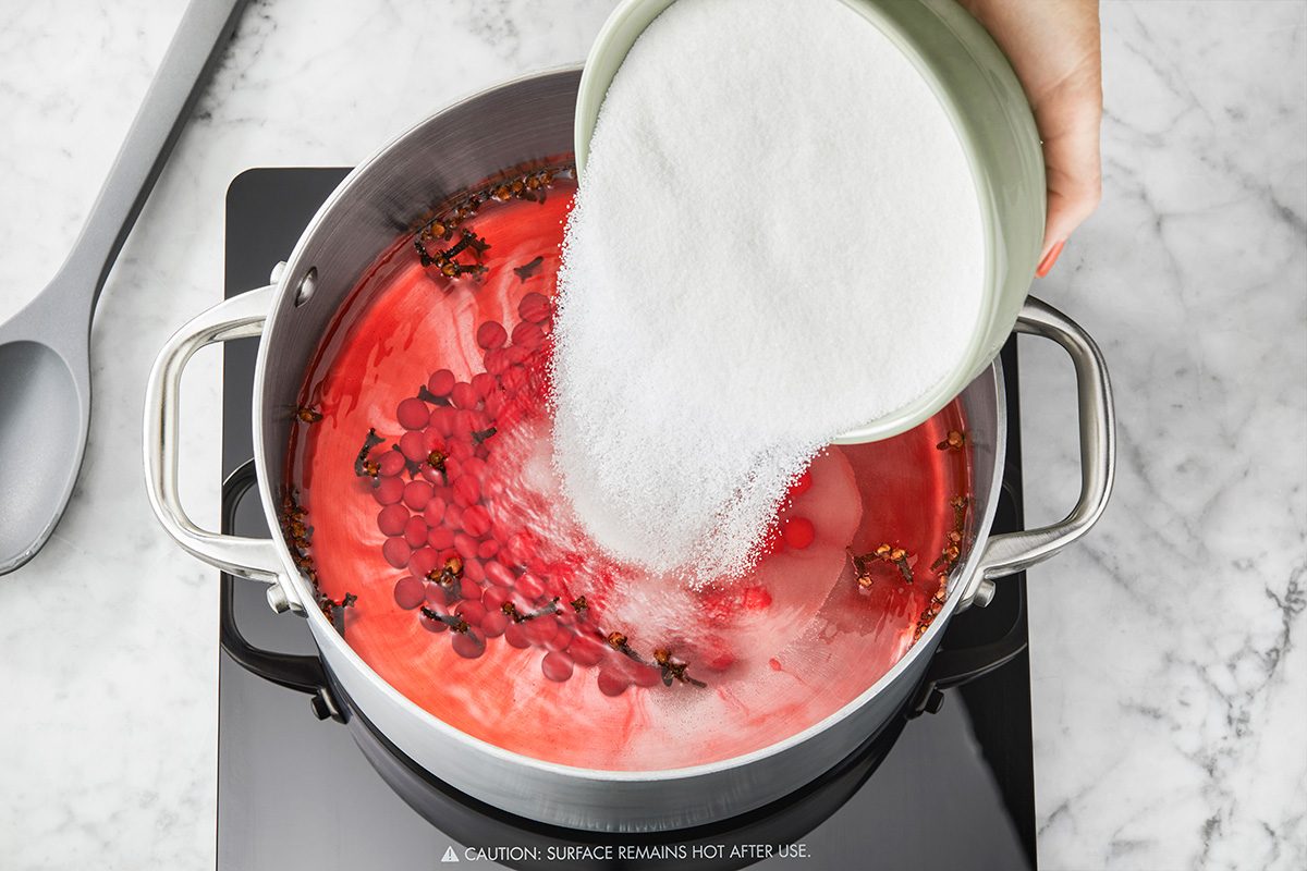 A hand pours a bowl of granulated sugar into a pot of red liquid with cranberries and spices, heating on a stovetop. A gray spoon rests nearby on a marble countertop.