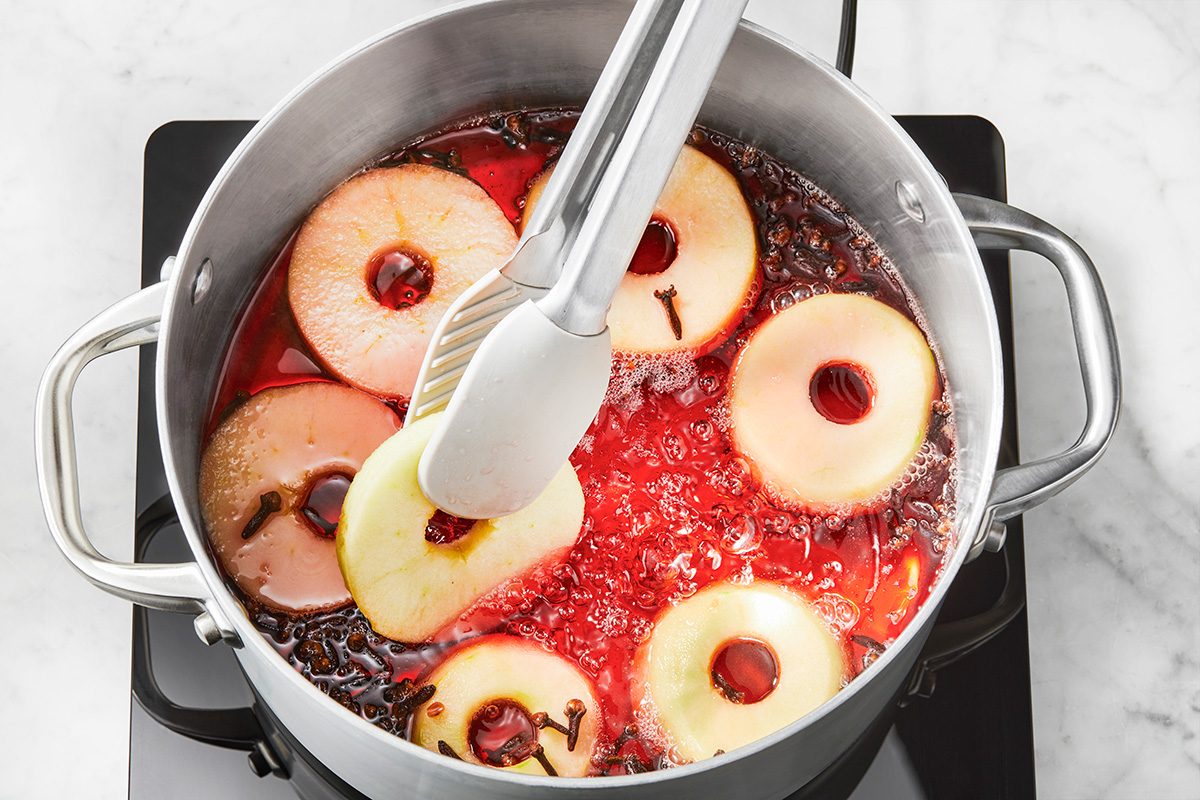 A pot on a stove contains round apple slices, cloves, and a red liquid, while metal tongs hold one apple slice above the simmering mixture.