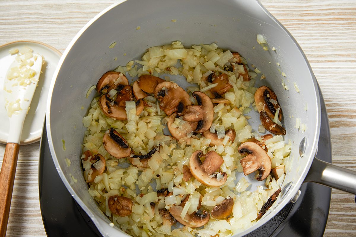 A saucepan on a stovetop containing chopped onions and sliced mushrooms being sautéed. A wooden spatula rests on the side of the pan. The background is a wooden surface.