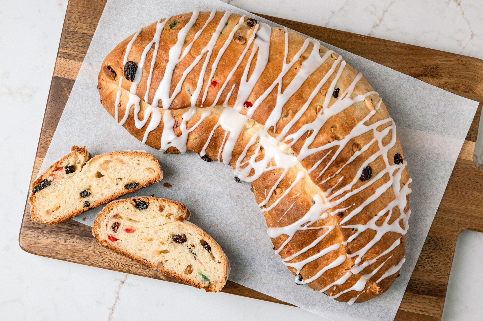 overhead shot of a large loaf of sweet bread displayed on a wooden cutting board, adorned with a glossy white icing drizzled in a decorative pattern across its surface; beside the loaf, two slices of the bread are laid out, the loaf is set on a piece of parchment paper