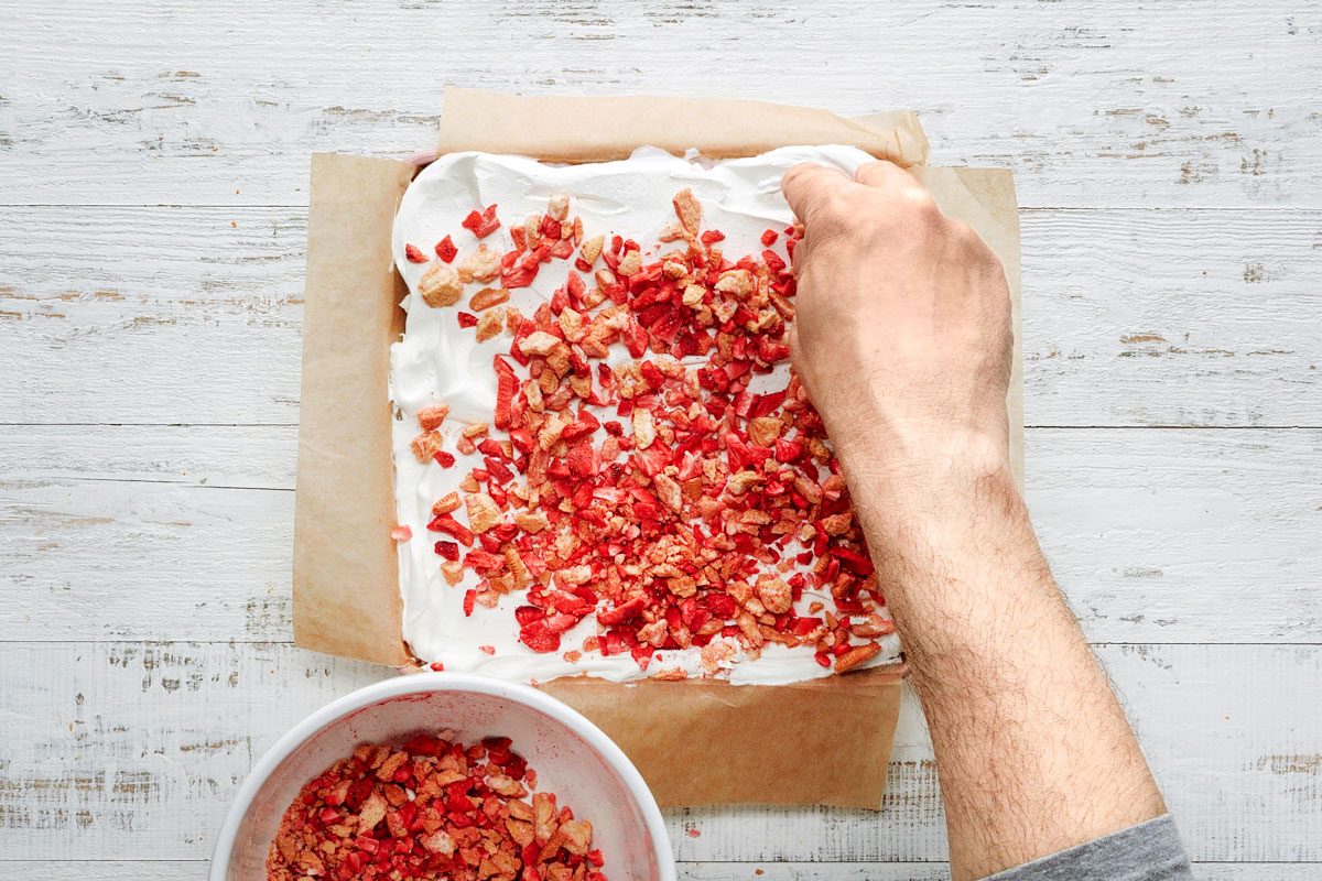 Overhead shot of Coarsely crush remaining cookies; Combine cookie crumbs and freeze-dried strawberries; sprinkle over whipped topping; Freeze; covered until firm; 8 hours or overnight; Remove cake from freezer; Lifting with parchment remove from pan; Gently peel off parchment; Let stand 10 minutes before cutting; garnish with fresh strawberries; white wooden surface;