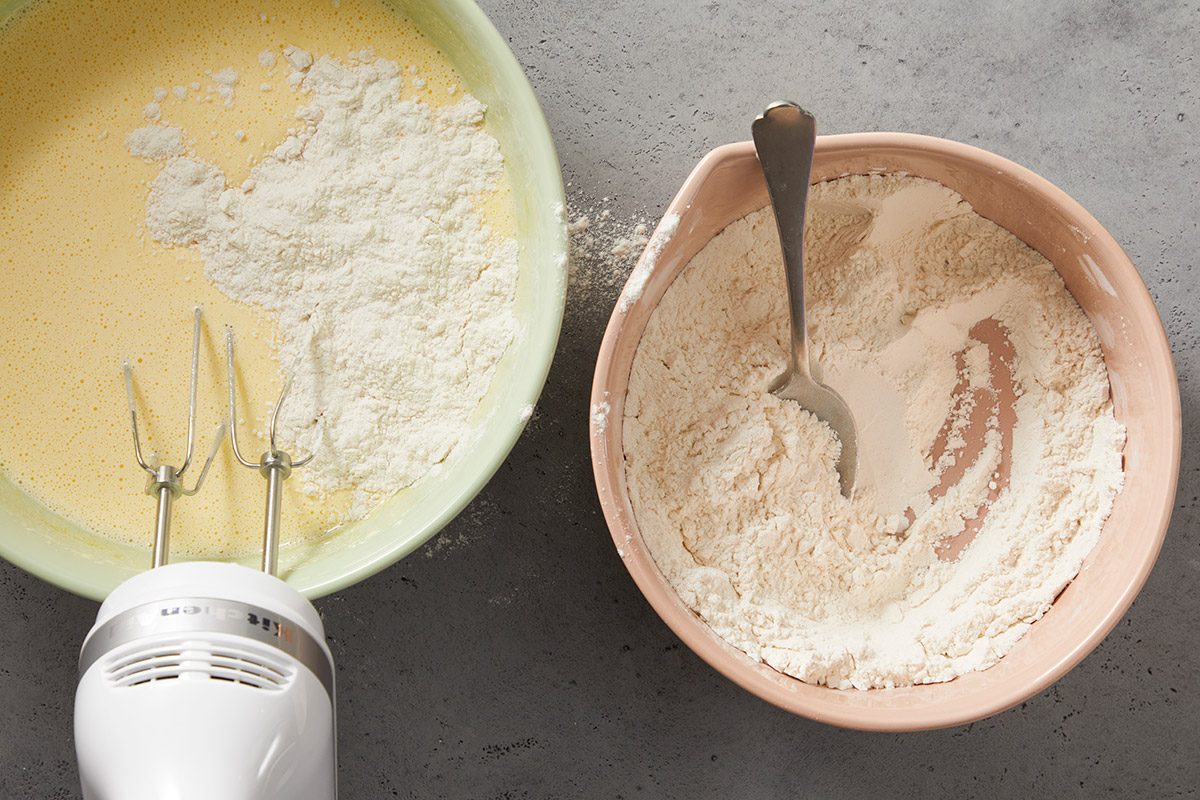 A hand mixer, a bowl with yellow batter and flour, and another bowl with flour and a spoon sit on a gray countertop, ready for baking preparation.