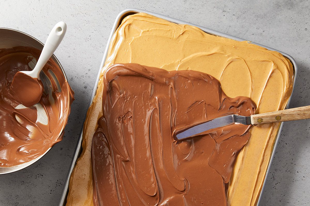 A baking tray with a layer of light brown peanut butter mixture being topped with melted chocolate, spread with an offset spatula. A mixing bowl with chocolate and a spatula sits nearby on a gray surface.