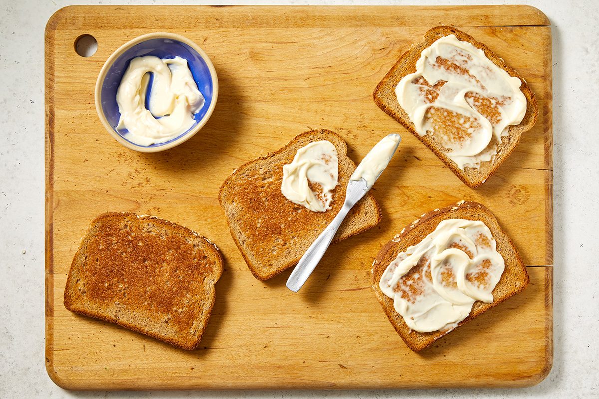A wooden cutting board holds four slices of toasted bread, some spread with butter or margarine. A butter knife and a small bowl of spread are also on the board.