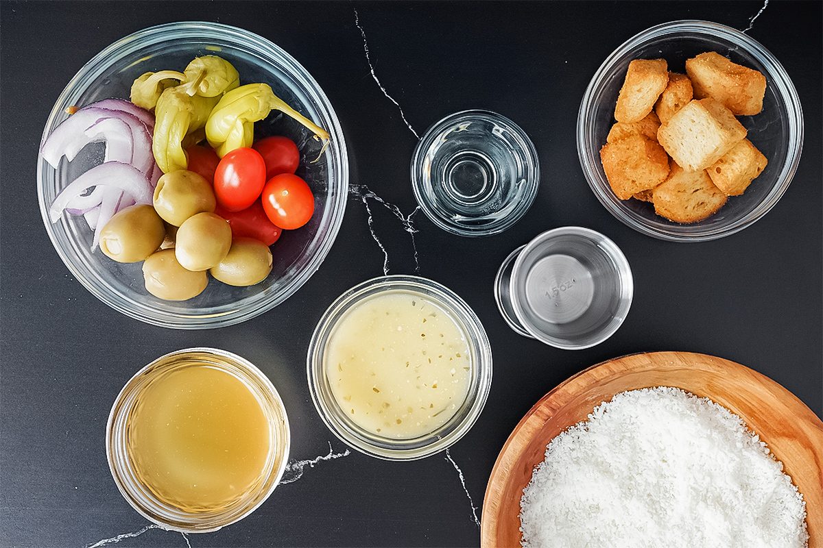 ViralaVarious ingredients in bowls on a dark surface, including sliced onions, olives, cherry tomatoes, dressing, croutons, and grated cheese. Two smaller bowls contain liquids.Olive Garden Salad Martini Lauren Habermehl For Taste Of Home Trends 02 Yvedit A