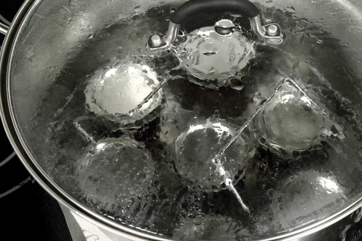 3/4th shot of a pot with four glass jars inside is on a stovetop; the pot is filled with boiling water; the water is bubbling and churning inside the pot; the jars are submerged in the water and the pot is covered with a lid; the lid is secured to the pot by a handle that has a large knob on top; there is a metal piece that is connected to the lid by a metal bar; the metal piece is also secured to the handle; the metal piece is used to hold the jars in place inside the pot; the whole pot is made of stainless steel and has a shiny finish;