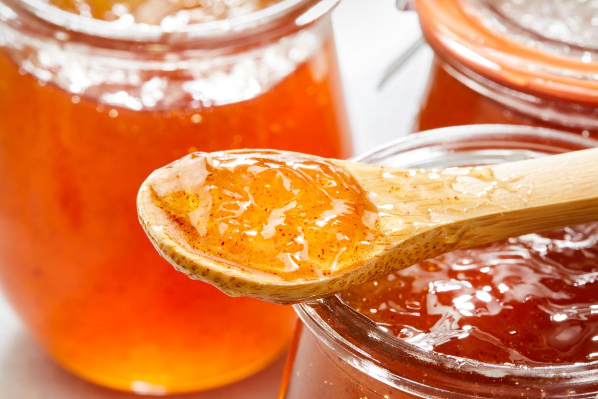 Close-up shot of Apple Cinnamon Jelly with a spoon resting over one jar; and additional jars in the background; all arranged on a marble surface;