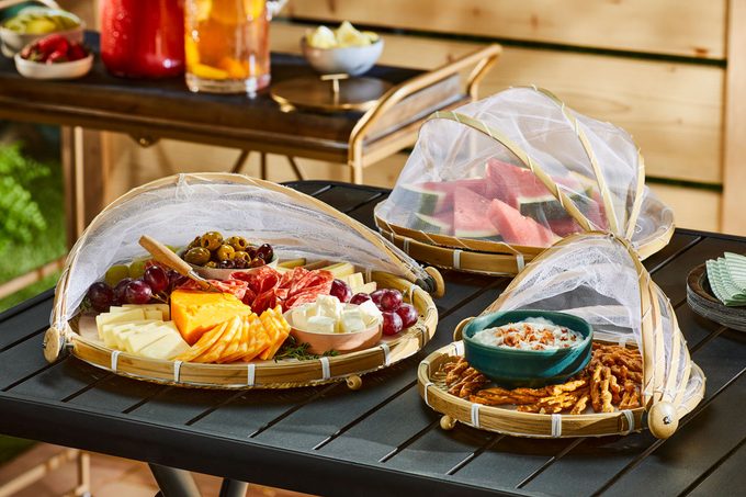 a beautifully arranged outdoor food spread on a dark table, with three circular platters featuring a variety of food items; the second platter holds slices of watermelon under a mesh cover; and the third platter is filled with pretzels alongside a bowl of dip, the food is neatly protected by mesh domes