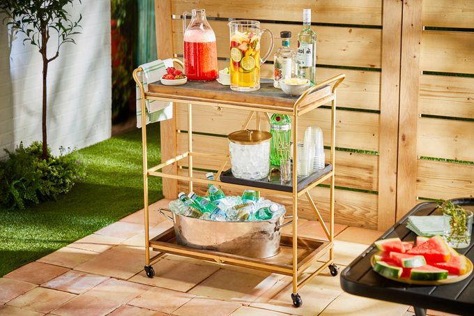 Drink cart set up in a backyard. Two batch drinks on the top shelf with garnishes, ice and cups on the second shelf, and additional bottled drinks in a silver tub on the bottom shelf. Watermelon on a table in foreground