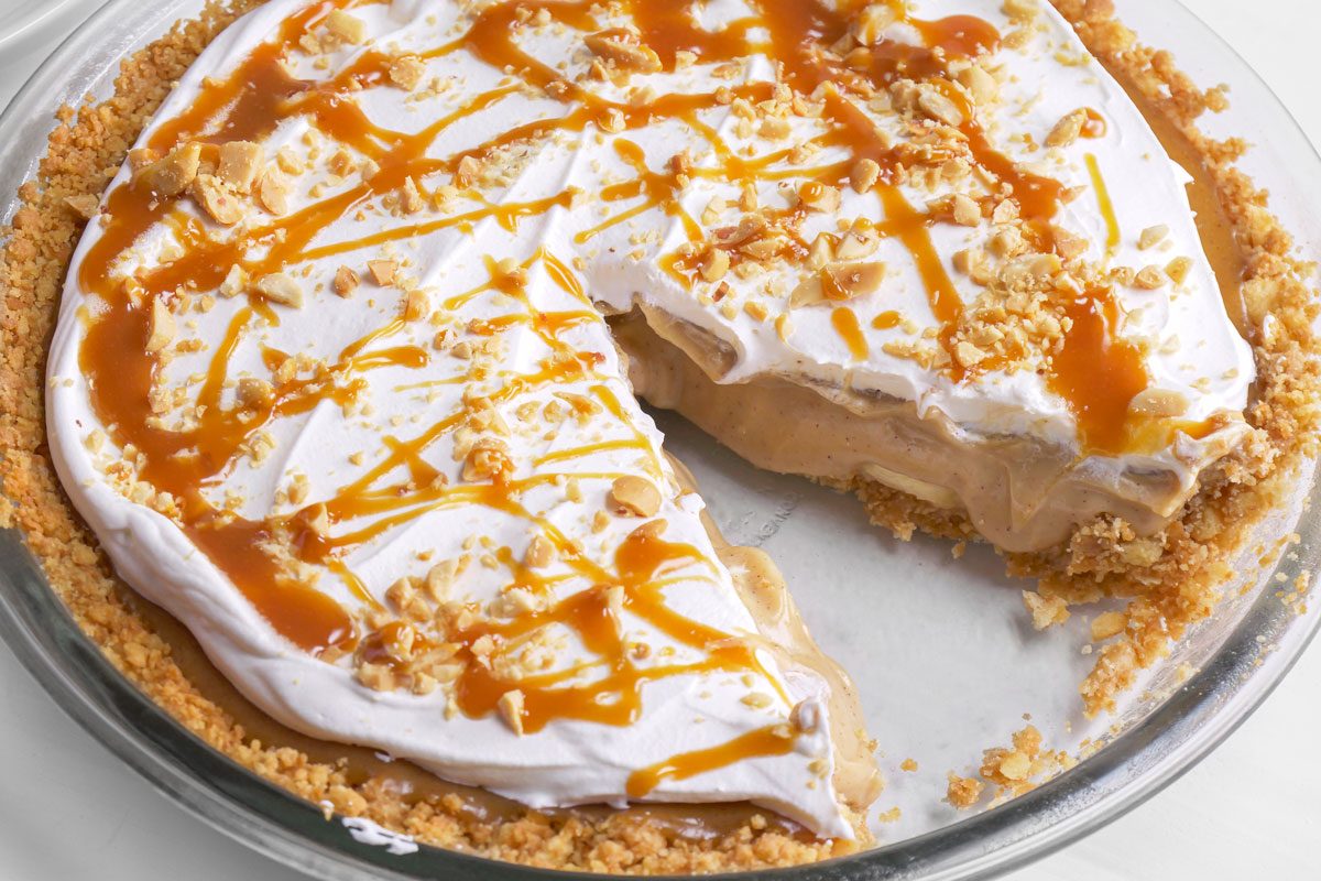 3/4th shot of A Bananas Foster creamy pie with a crumb crust, topped with whipped cream, chopped nuts, and drizzled caramel sauce. A large slice has been removed, showing the pie’s smooth filling inside a glass pie dish