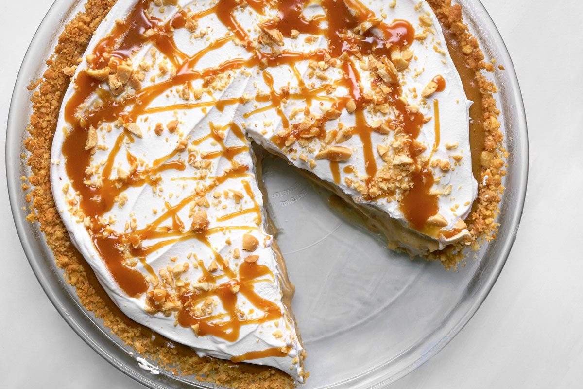 overhead shot of A Bananas Foster pie with a crumbly crust, topped with whipped cream, caramel drizzle, and crushed nuts, sits in a glass pie dish with one large slice missing