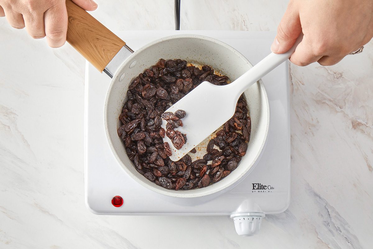 overhead shot of A person stiring a pan of raisins with a white spatula on a white electric hot plate, the background is white