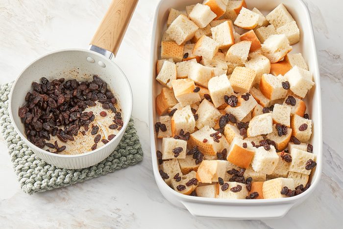 3/4th shot of a white baking dish filled with cubed bread and scattered raisins sits next to a small saucepan with cooked raisins on a knitted trivet, on a light marble surface