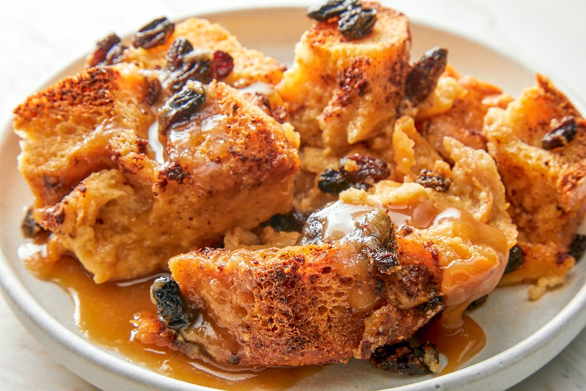 closeup shot of a plate of Bread Pudding With Rum Sauce topped with raisins and drizzled with caramel sauce, served on a light colored dish