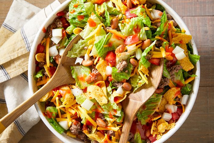 A large bowl of taco salad with lettuce, diced tomatoes, onions, shredded cheddar cheese, ground beef, pinto beans, and corn chips, tossed together with two wooden salad servers.
