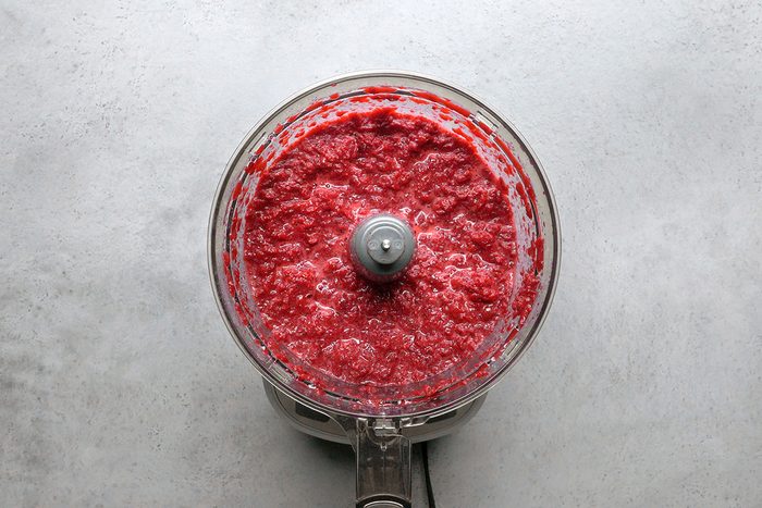 A food processor filled with blended red berry puree sits on a light gray countertop, viewed from above.
