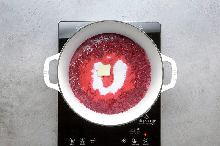 A white pot on a black stovetop contains a red mixture with a pat of butter and a heap of sugar in the center, ready to be mixed.