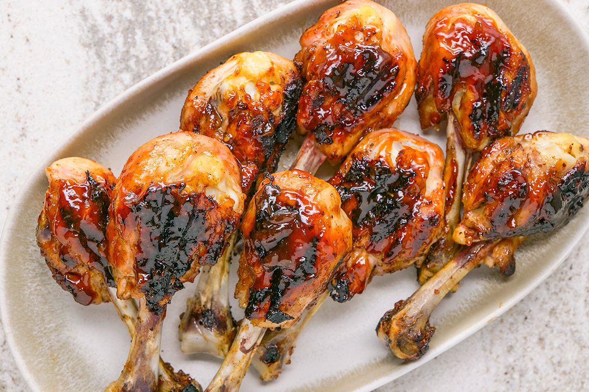 Seven grilled chicken drumsticks glazed with a shiny, dark barbecue sauce are arranged on a light oval plate, showing caramelized and charred spots on the skin.