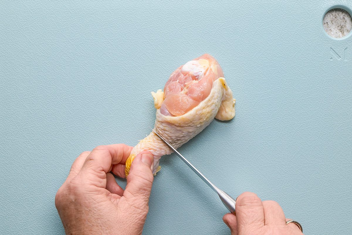 A person uses a knife to cut a raw chicken drumstick on a light blue cutting board.