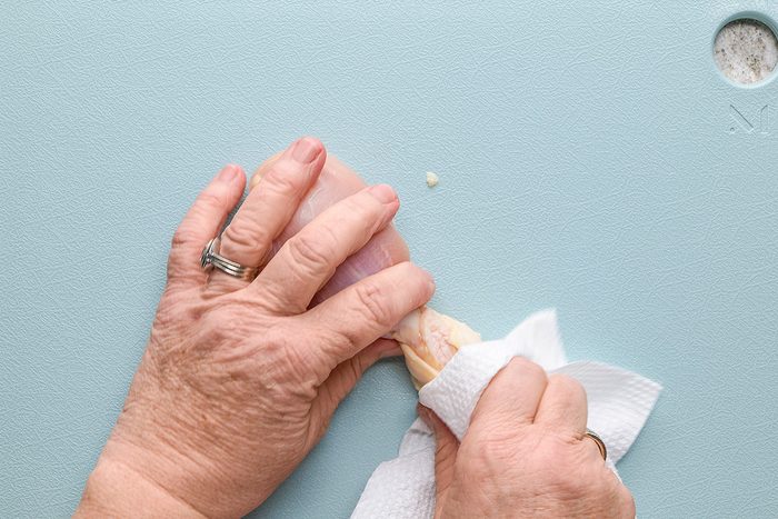 A person using a paper towel to grip and remove the skin from a raw chicken drumstick on a light blue cutting board.