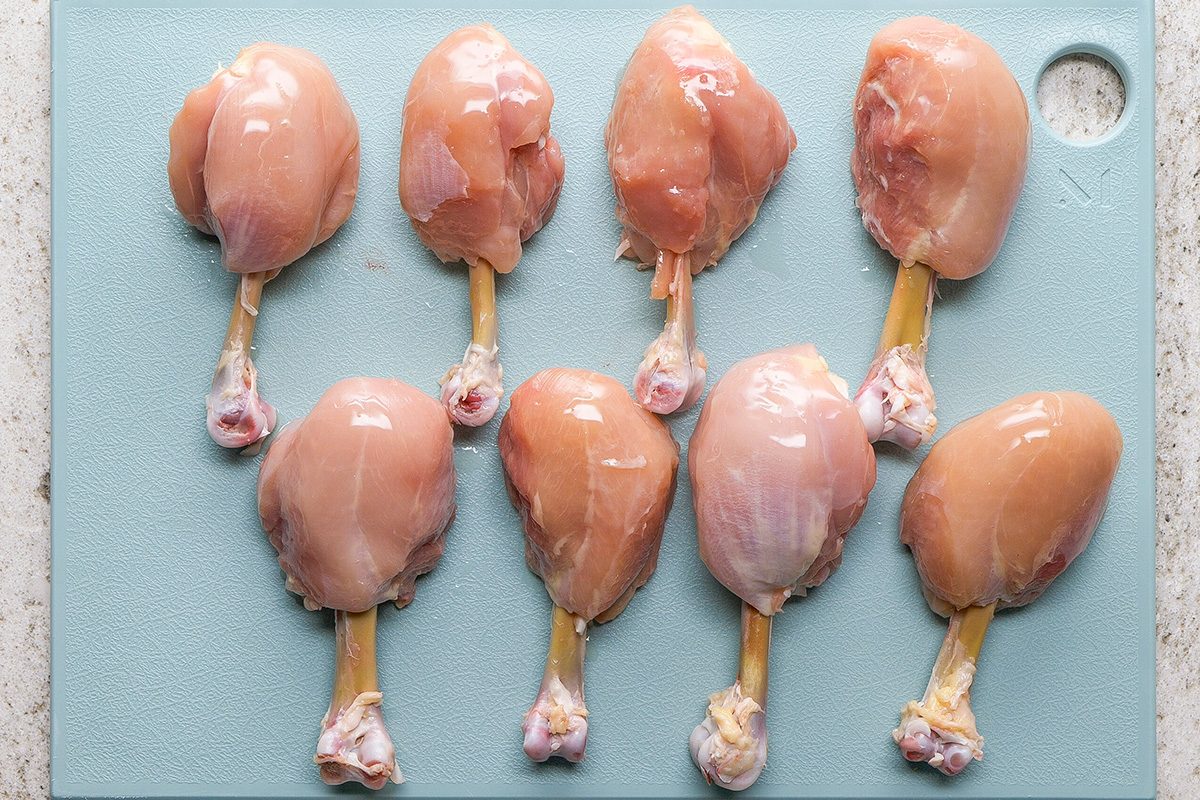 Eight raw chicken drumsticks with the meat pushed up to one end are arranged on a light blue cutting board, ready to be cooked or seasoned.