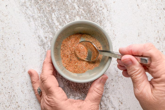 A person holds a small bowl of blended spices in one hand and stirs the mixture with a spoon using the other hand. The bowl is on a light-colored countertop.