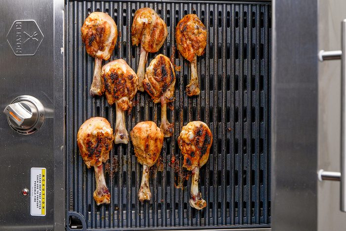 Grilled chicken drumsticks cooking on an outdoor barbecue grill, showing char marks on the meat. The grill has metal grates and a temperature control knob on the left side.