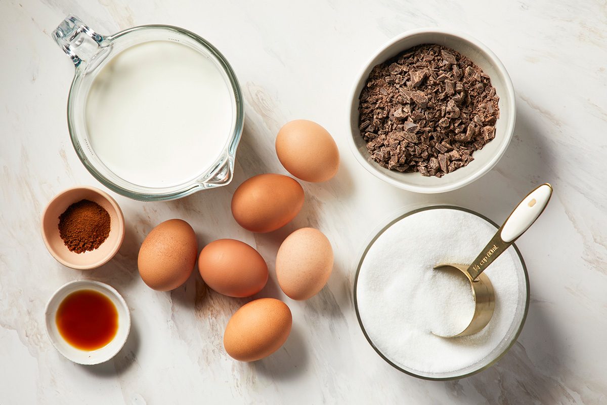 A top view of baking ingredients on a white surface, including eggs, a cup of milk, a bowl of cocoa powder, a bowl of sugar with a measuring cup, vanilla extract, and ground cinnamon.