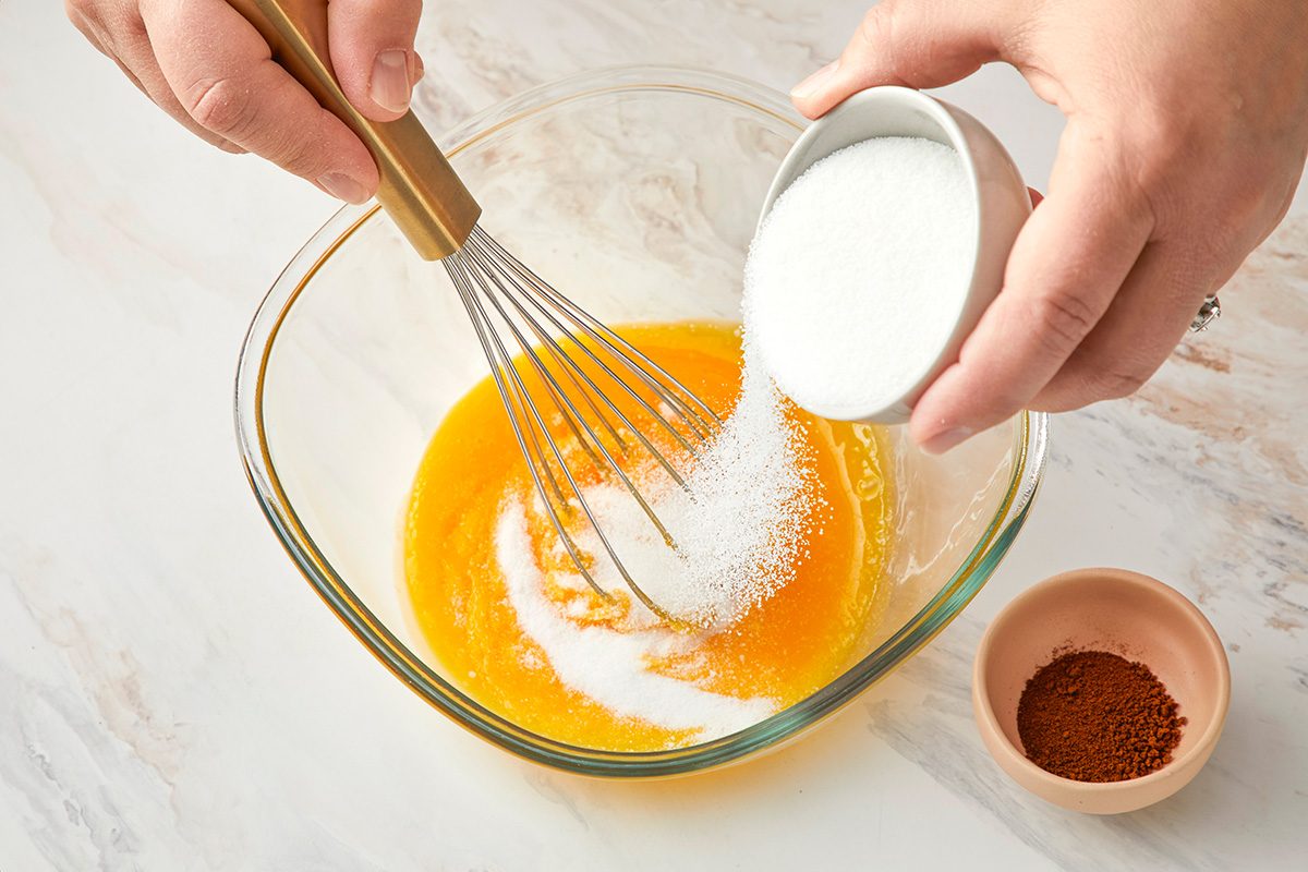 A person adds sugar to a glass bowl containing whisked eggs while holding a whisk; a small bowl of ground spice sits nearby on a marble surface.
