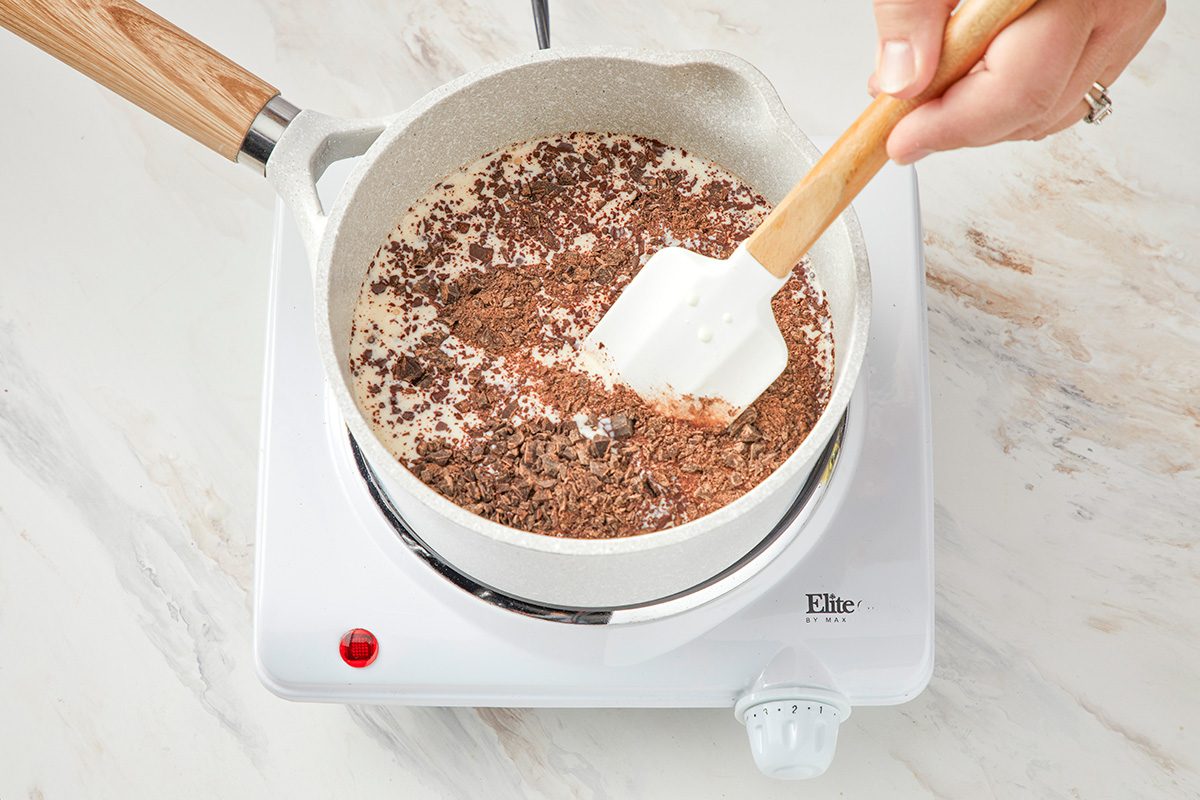 A hand stirs chocolate shavings into milk in a white saucepan on a small electric stovetop with a white marble background.