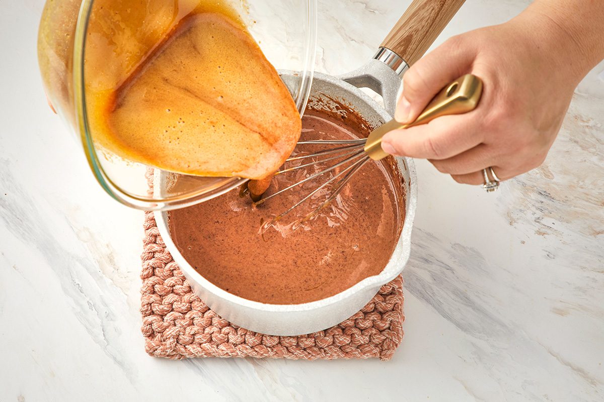 A hand whisks chocolate batter in a white bowl while another hand pours beaten eggs from a glass bowl into the mixture on a marble countertop.
