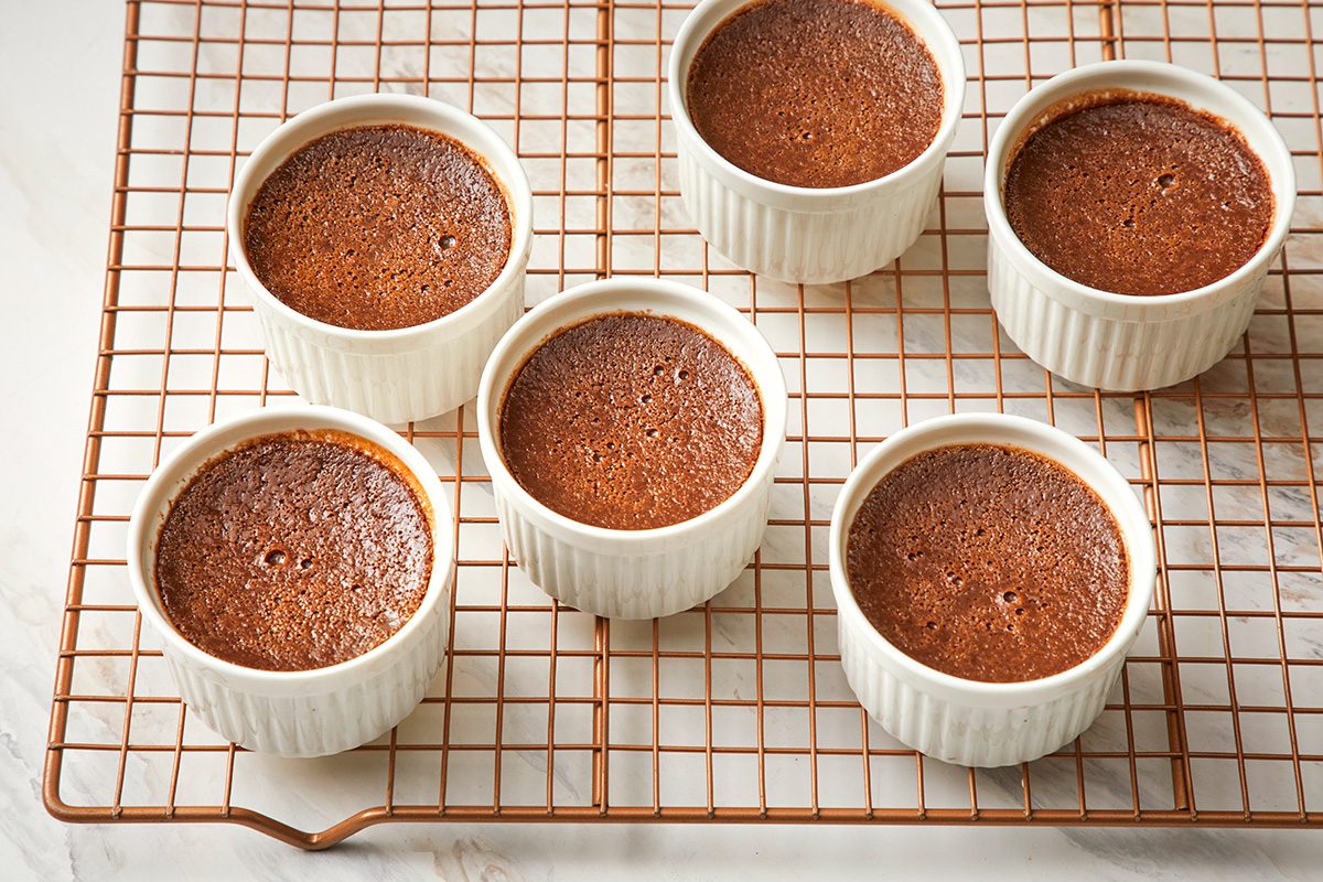 Six chocolate soufflés in white ramekins are cooling on a wire rack, seen from above on a light surface.