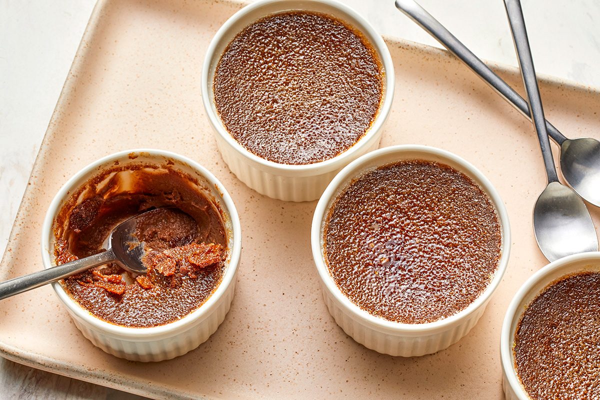 Four ramekins of chocolate crème brûlée are on a tray, with one partially eaten to reveal a creamy interior. Three metal spoons are placed nearby on the tray.