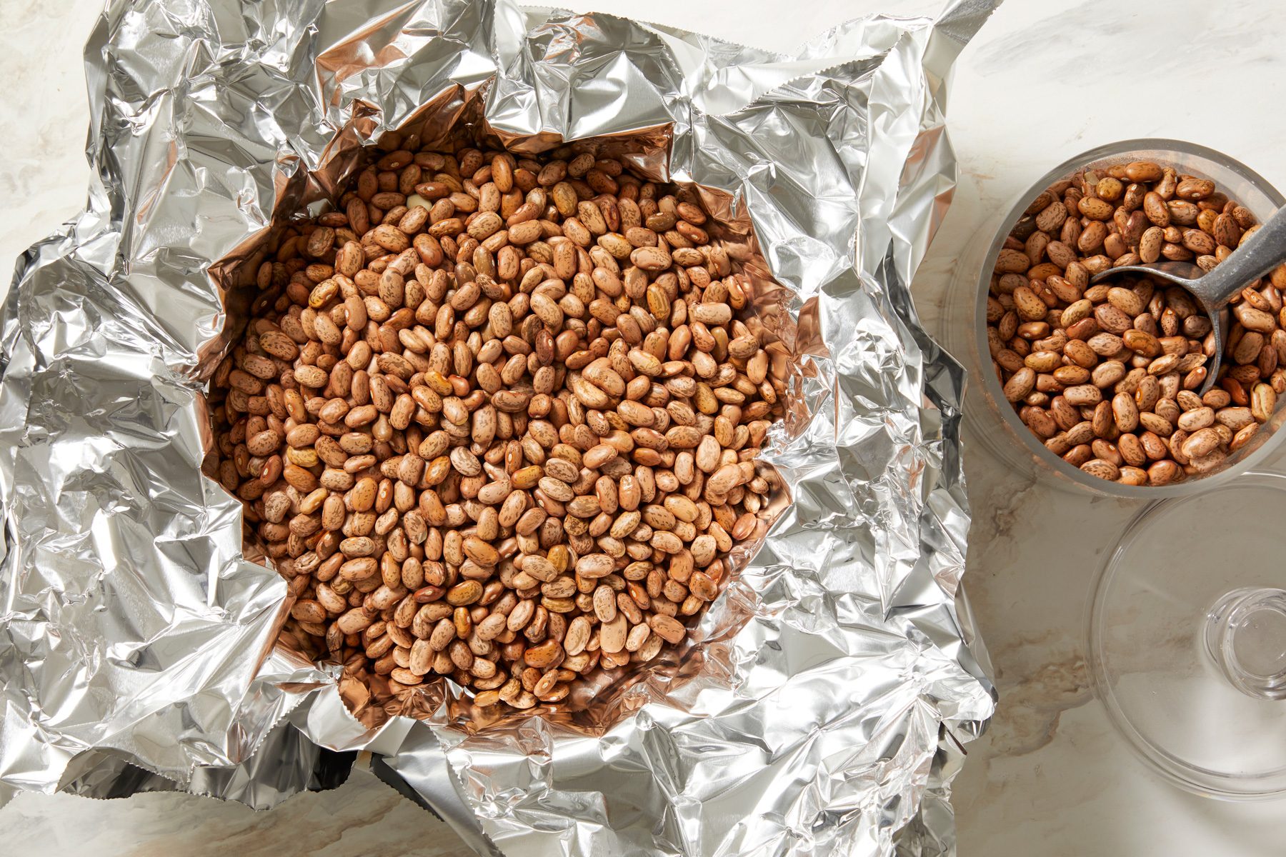 overhead shot of a large mound of dried beans nestled within aluminum foil; next to the foil, there is a smaller glass bowl containing similar beans, also featuring a metal spoon resting on top; the background is a subtle marbled surface