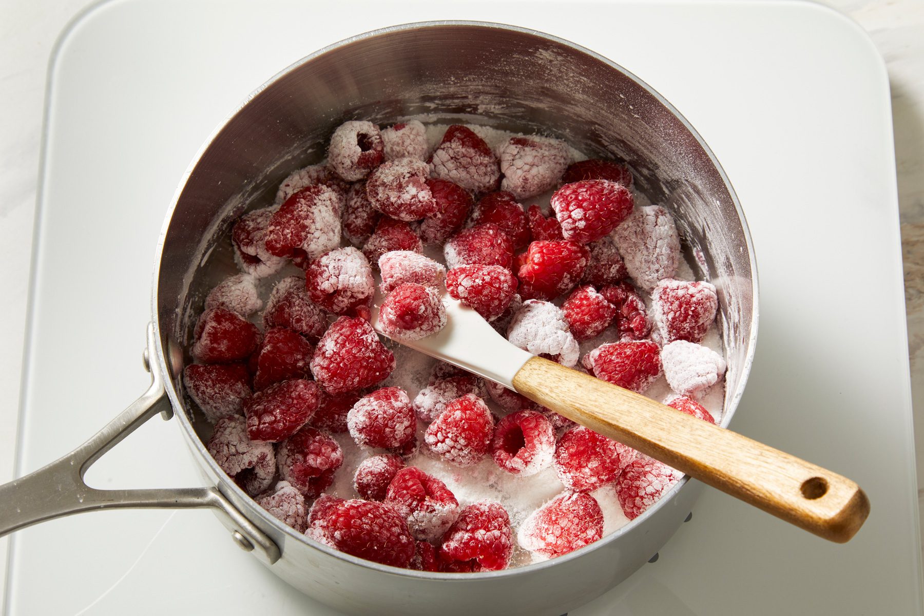 3/4th shot of a pot filled with fresh raspberries is visible; the raspberries are coated with a layer of sugar, a white spatula with a wooden handle rests among the berries; the background is a plain white surface