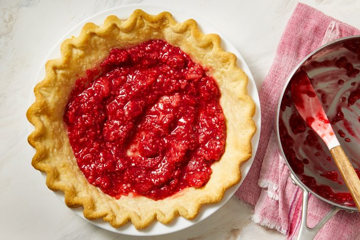 overhead shot of a pie crust that is partially filled with red raspberry filling; beside the pie crust, there is a metallic bowl containing some of the raspberry filling, along with a spatula coated in the same filling; a pink and white striped cloth is placed underneath the bowl