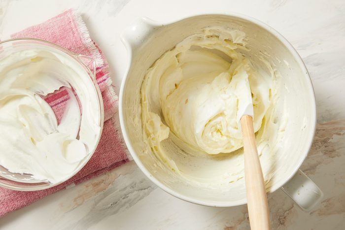 overhead shot of two bowls of creamy mixtures on a light, textured countertop; on the right, a larger bowl contains whipped cream or frosting, with a wooden spatula resting in it; on the left, a smaller glass bowl is holding similar cream, while there is pink dish towel underneath