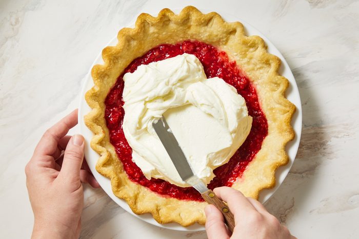 overhead shot of pie sitting in a white dish with a crimped edge; a hand is seen holding a spatula, which is being used to spread the creamy topping over the red filling; the background is a light marbled surface