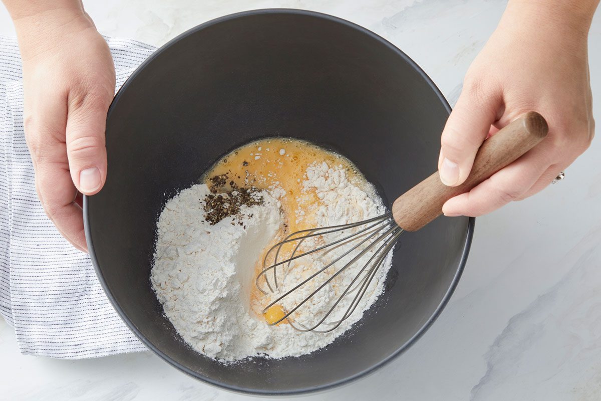 A person holds a black mixing bowl with flour, cracked egg, and pepper, whisking the ingredients together on a light-colored surface.